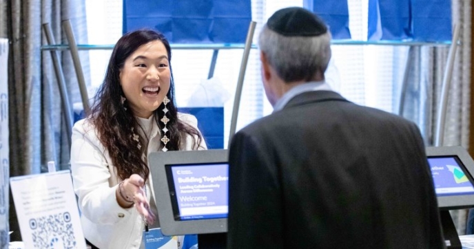 Smiling woman at registration booth for conference talking to Council member, man wearing a yarmulke